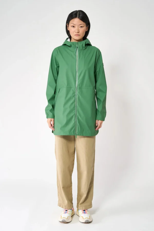 Una modella indossa la giacca impermeabile verde Comfrey di Tanta Rainwear, chiusa sul davanti, abbinata a pantaloni beige larghi e sneakers colorate.