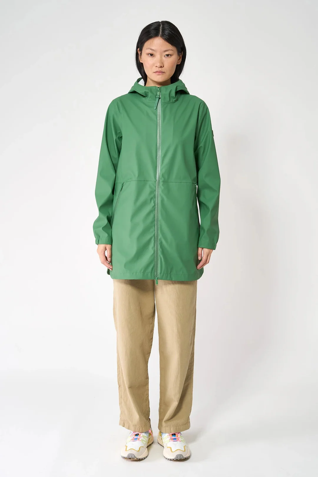Una modella indossa la giacca impermeabile verde Comfrey di Tanta Rainwear, chiusa sul davanti, abbinata a pantaloni beige larghi e sneakers colorate.