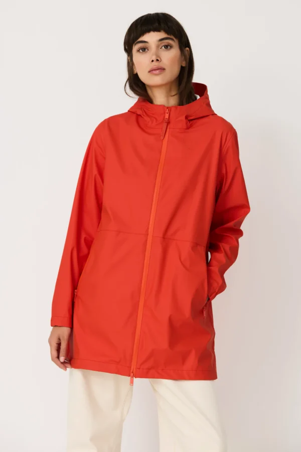 Una modella indossa la giacca impermeabile rosso vivo Grenadine di Tanta Rainwear, con cappuccio e mani nelle tasche, abbinata a pantaloni bianchi larghi.