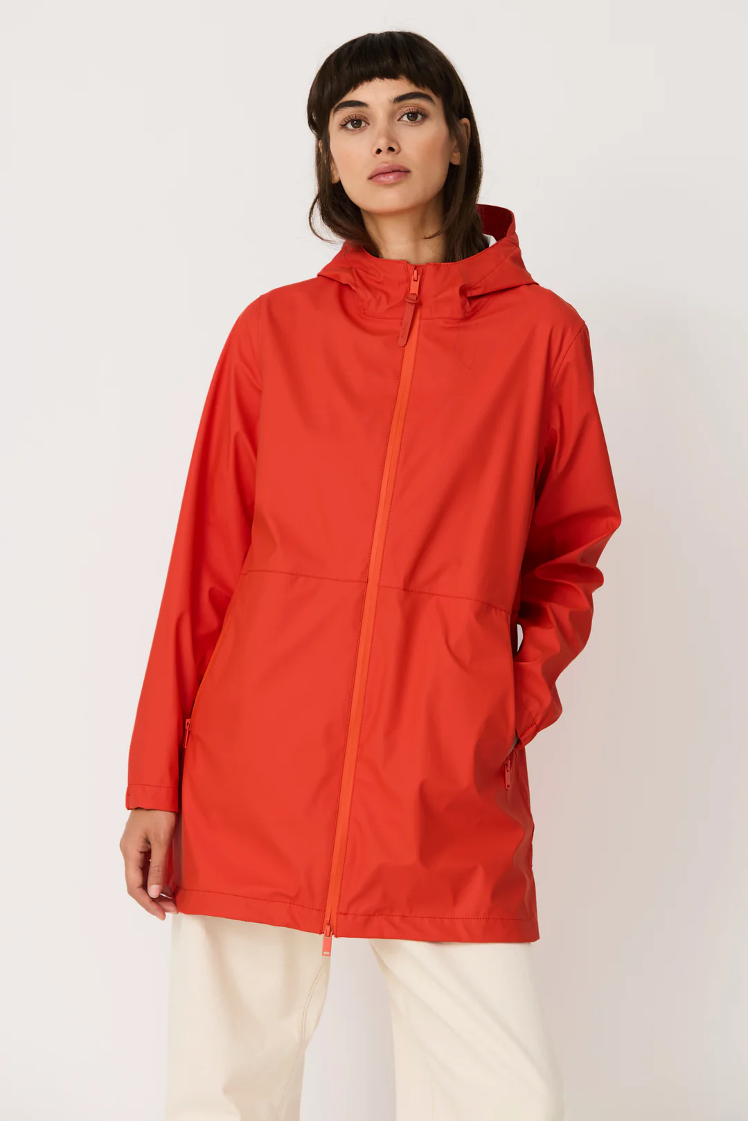 Una modella indossa la giacca impermeabile rosso vivo Grenadine di Tanta Rainwear, con cappuccio e mani nelle tasche, abbinata a pantaloni bianchi larghi.