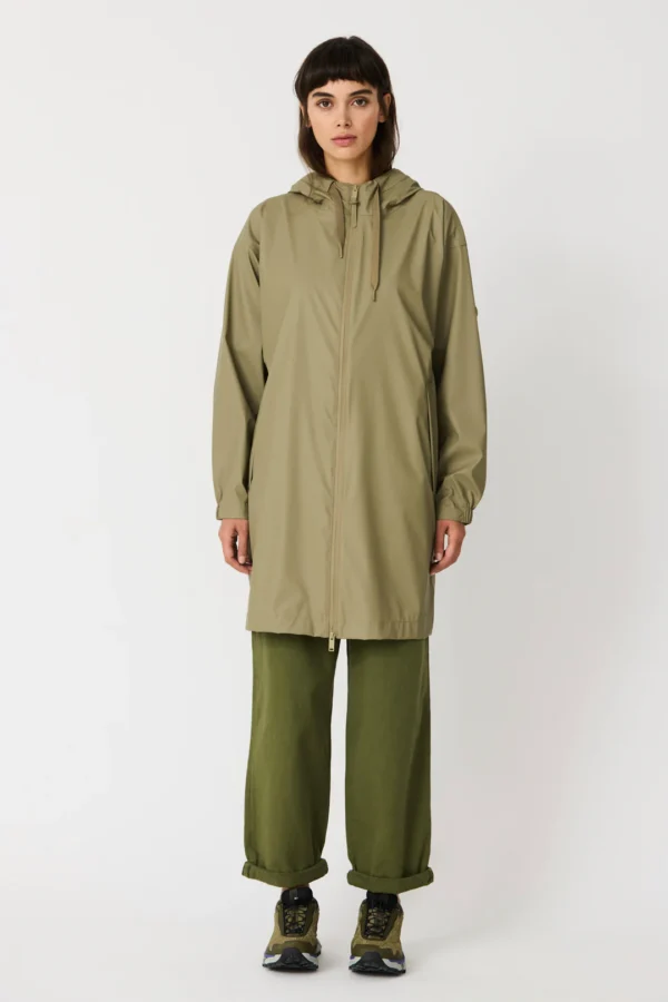 Una modella indossa la giacca impermeabile verde Mermaid/Ondina di Tanta Rainwear, più lunga e con cappuccio, abbinata a pantaloni verde oliva larghi e scarpe da trekking.