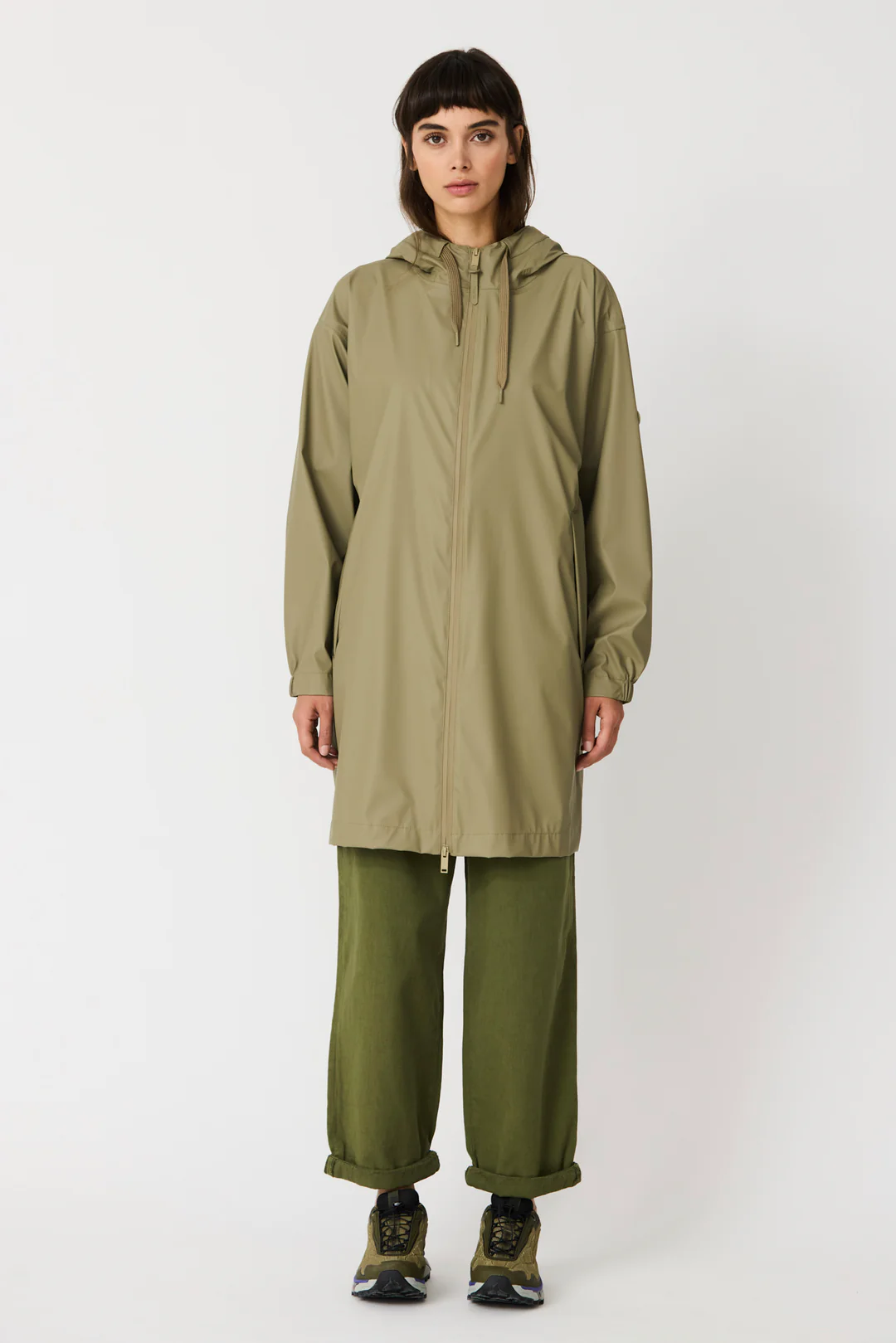 Una modella indossa la giacca impermeabile verde Mermaid/Ondina di Tanta Rainwear, più lunga e con cappuccio, abbinata a pantaloni verde oliva larghi e scarpe da trekking.