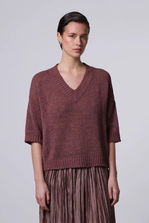 Un primo piano di una modella con capelli corti che indossa una maglia a maglia grossa, boxy e a maniche corte, color bordeaux, di Roberto Collina, con scollo a V.
