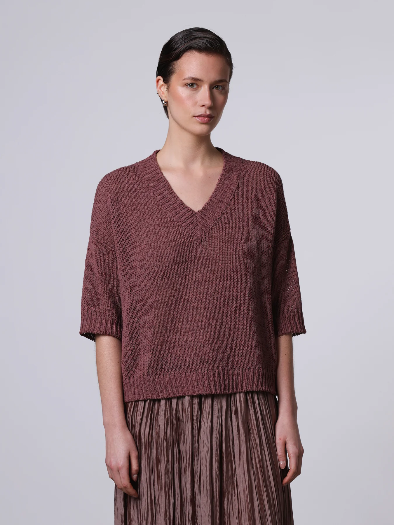 Un primo piano di una modella con capelli corti che indossa una maglia a maglia grossa, boxy e a maniche corte, color bordeaux, di Roberto Collina, con scollo a V.